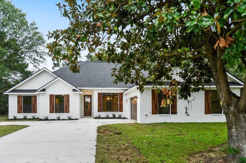 White brick house with brown shutters and a concrete driveway, fronted by green grass and a tree.