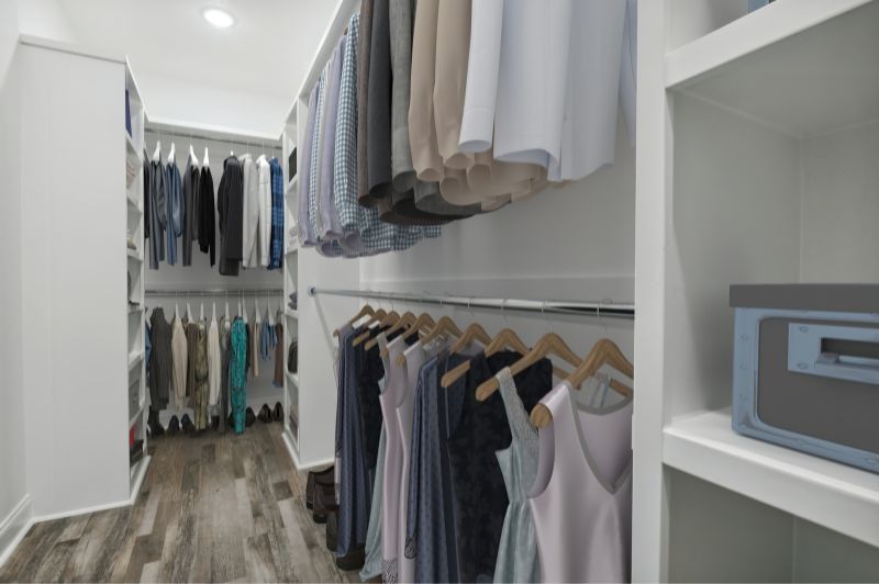 Walk-in closet with clothes hanging on rods. White shelves, wooden floors, and a blue box visible.