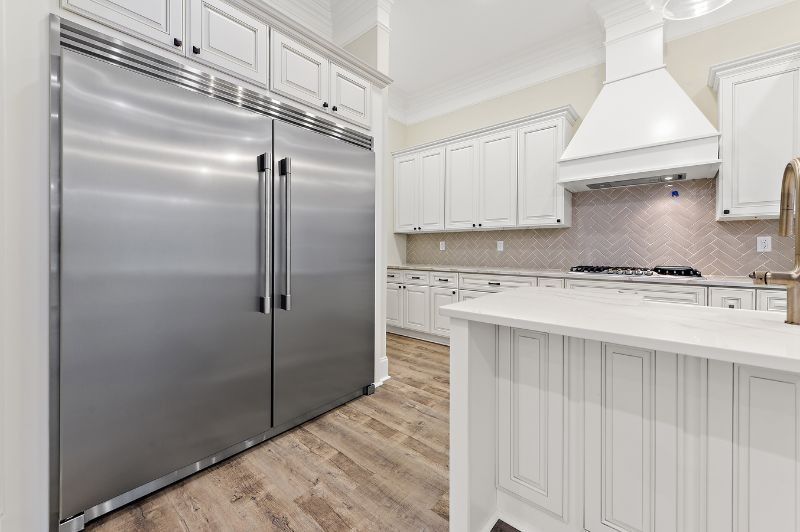 Stainless steel refrigerator in white kitchen with white cabinets and light wood flooring.