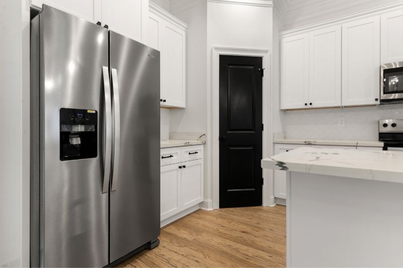 Stainless steel refrigerator in a bright kitchen with white cabinets and a black door.
