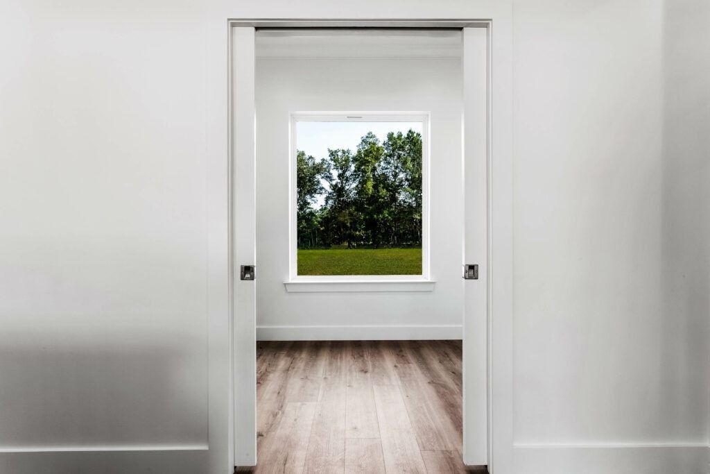 A white doorway frames a view of a window overlooking green trees and grass; room has wood flooring.