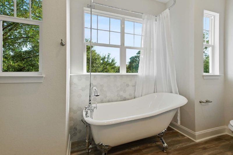 Claw-foot bathtub by a window with a view of trees, in a bathroom with white walls and a wood-look floor.