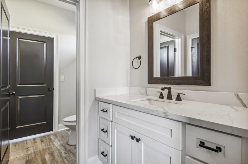White bathroom with marble countertop, dark wood framed mirror, and dark door.