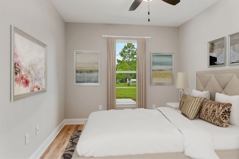 Bedroom with neutral tones, a bed with pillows, art, and a window with a view of greenery.
