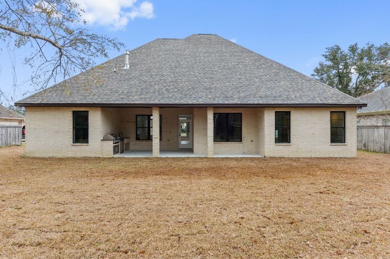 Back of a light brick house with a covered patio, windows, and a brown lawn under a blue sky.