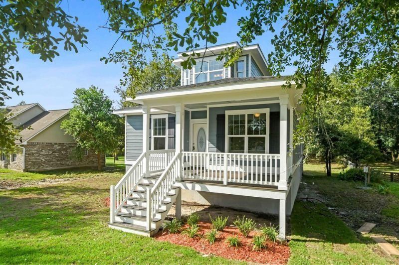 Blue cottage with white porch and stairs, nestled among green trees.