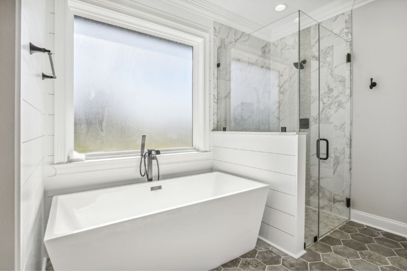 Modern white bathroom with a soaking tub, walk-in shower, gray hexagon tile floor, and frosted window.