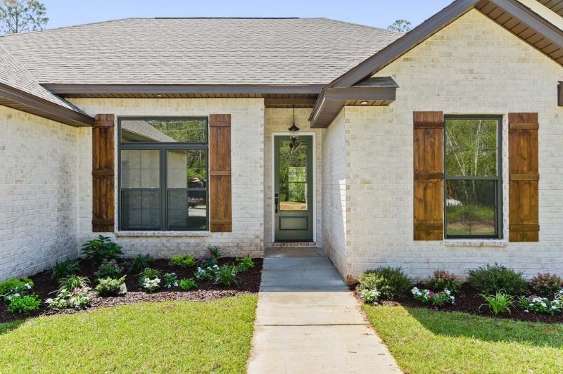 Beige brick house with brown shutters, windows, and door frame. Green lawn and landscaping.