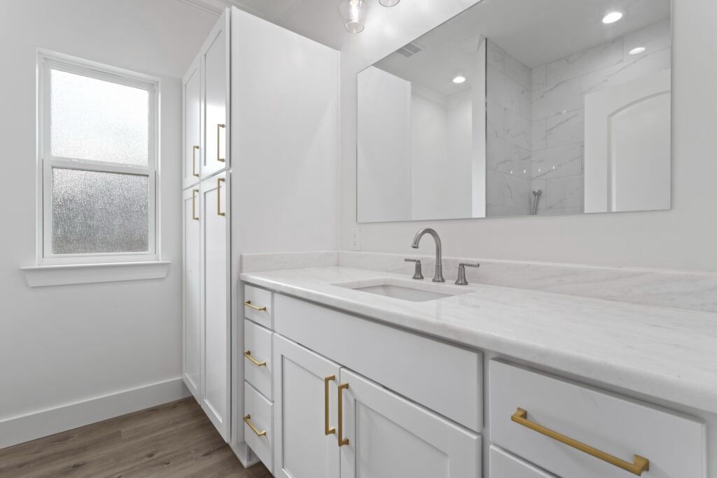 White bathroom with vanity, tall cabinet, window, and mirror; gold hardware.