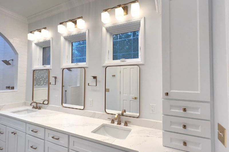 Bright bathroom with three mirrors, sinks, white cabinets, and brass fixtures.