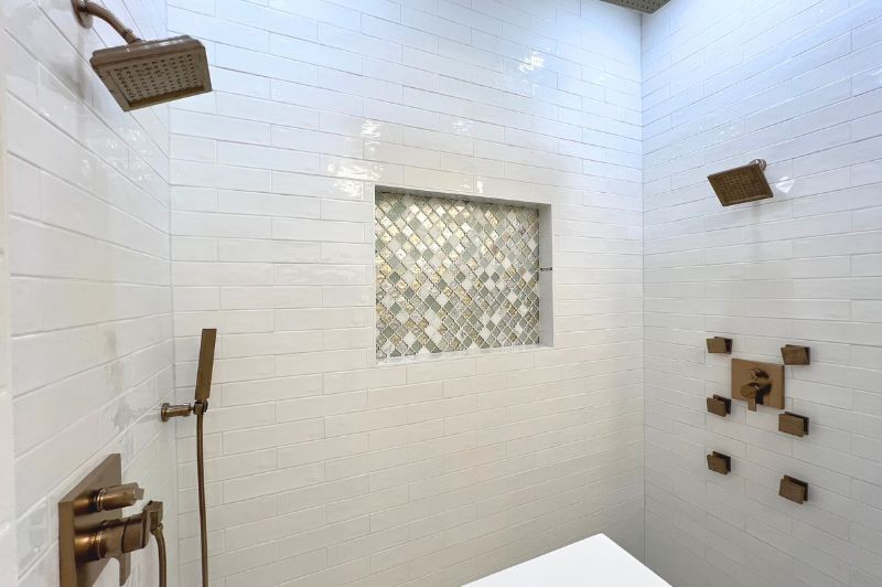 Modern white tiled shower with bronze fixtures, a tiled niche, and multiple shower heads.