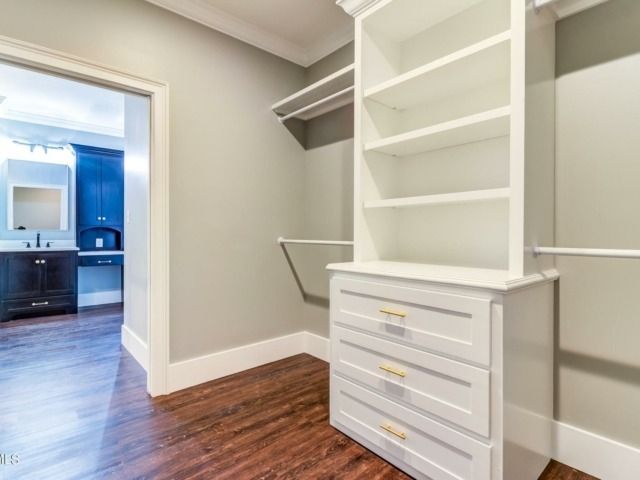 Walk-in closet with white built-ins, drawers, and shelves. Dark wood floor, gray walls, and an open doorway to a bathroom.