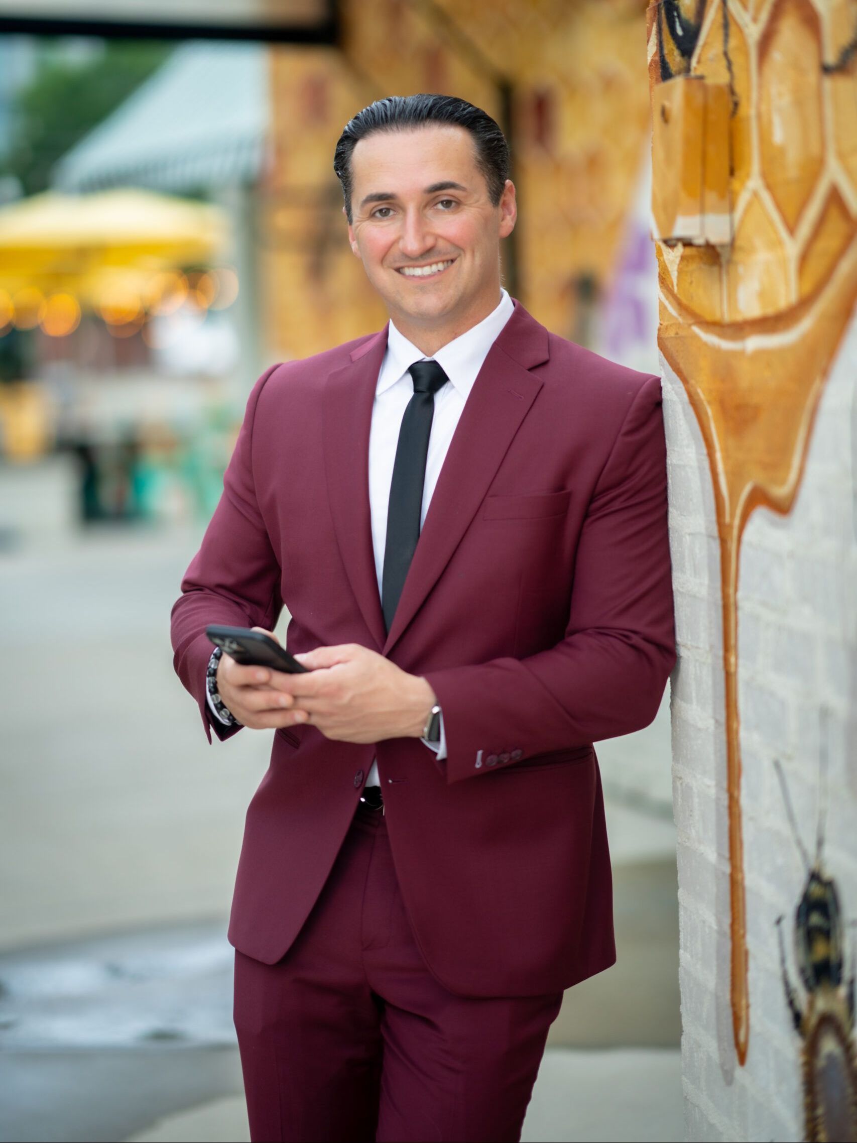 Mitch Boraski in a maroon suit and tie is leaning against a wall while holding a cell phone.