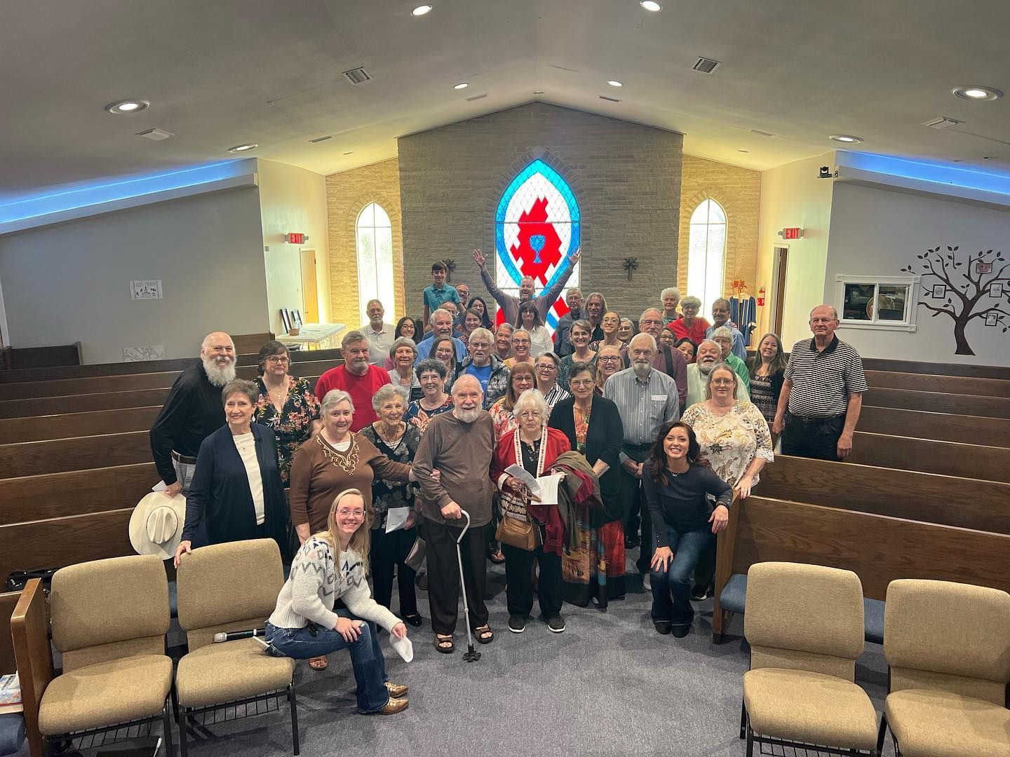 Group of people in a church sanctuary, posing for a photo in front of the altar with a stained glass window.