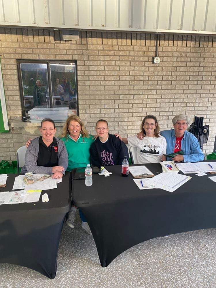 Five people smiling at a table, likely at an outdoor event. Brick wall background.