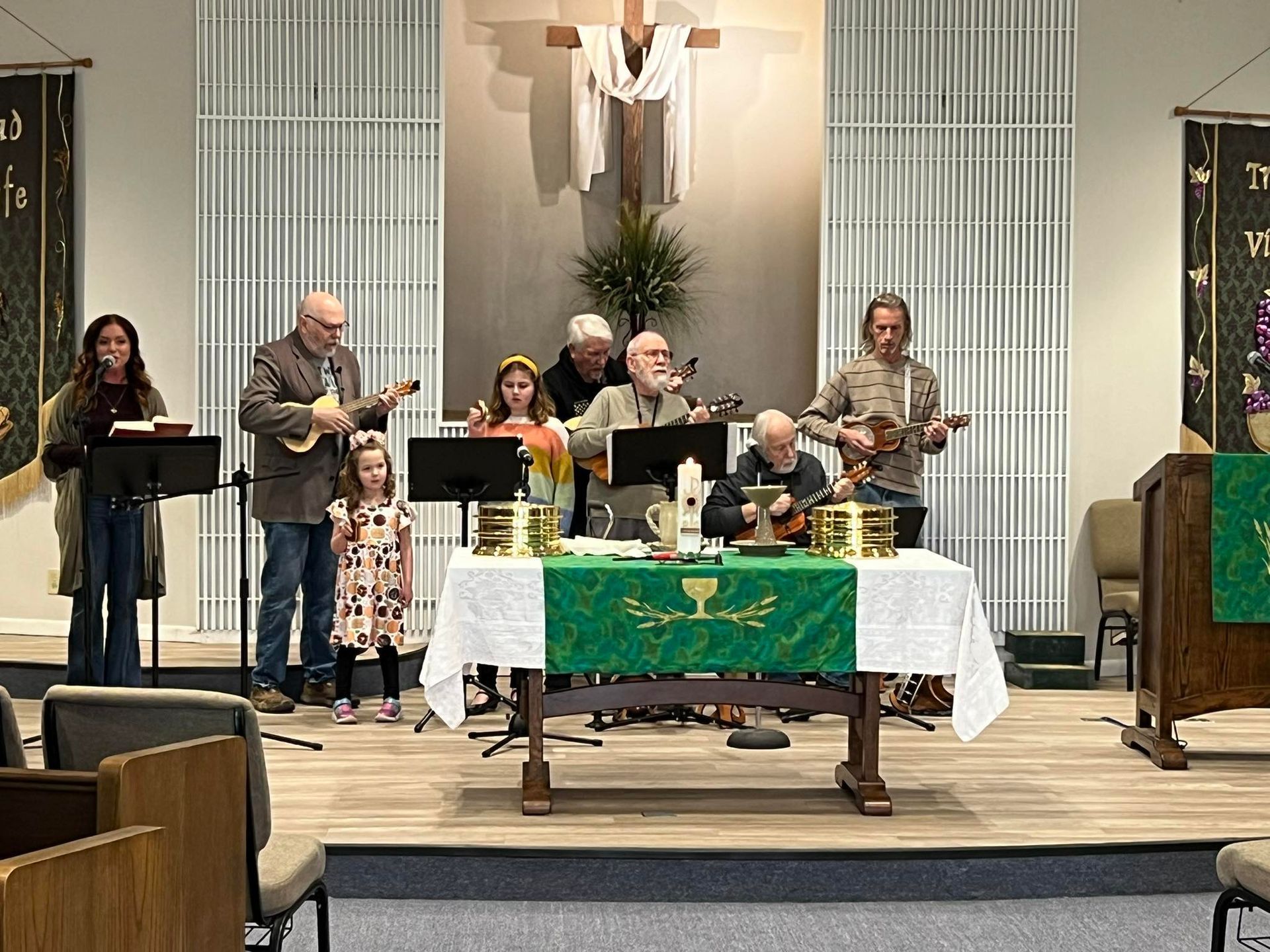 Worship band performing on a stage in a church. Musicians play instruments, sing, and stand before a table with a cross.