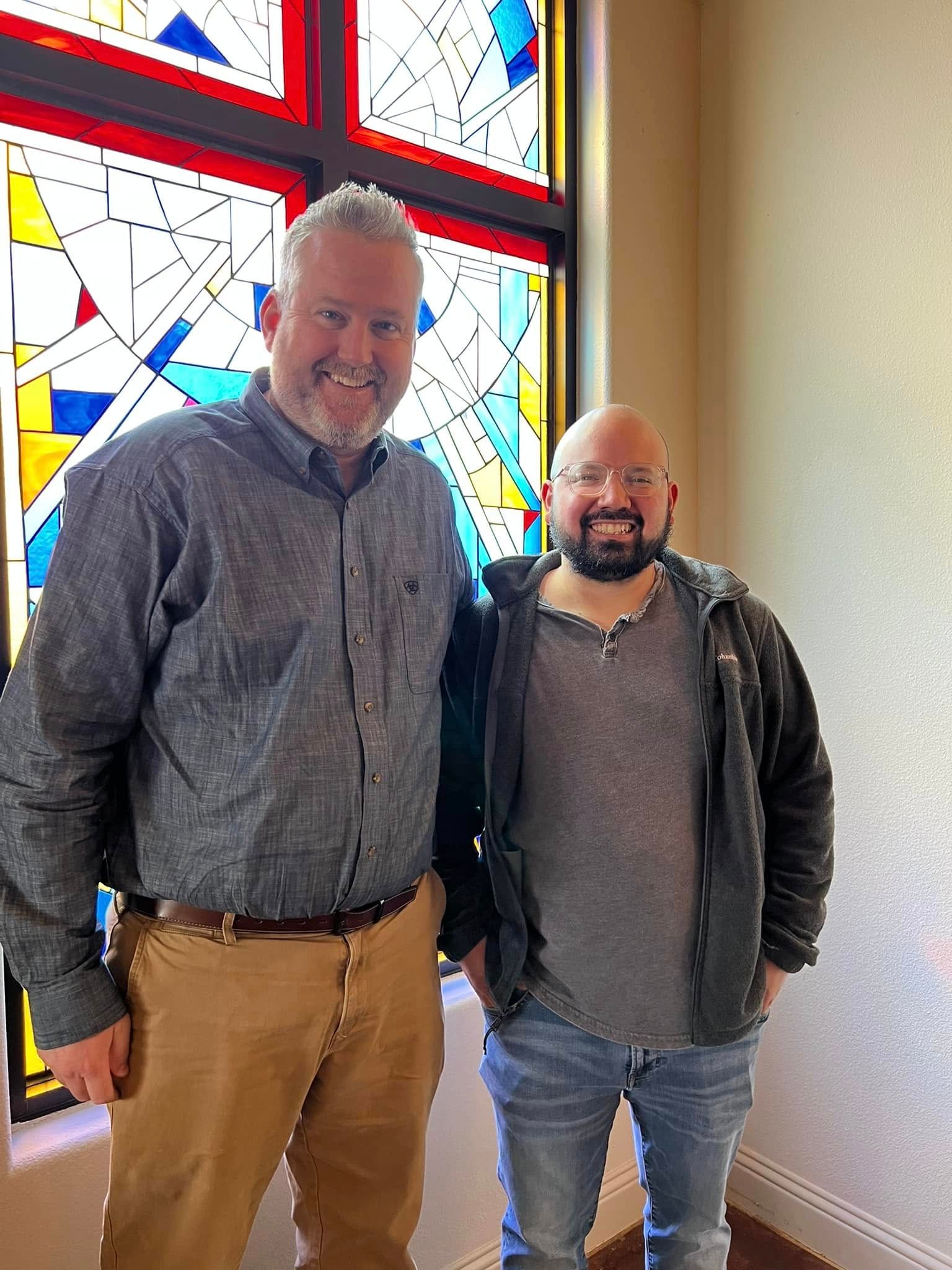 Two men smiling, posing by a colorful stained glass window. One wears a blue shirt, khaki pants, the other a hoodie, jeans.