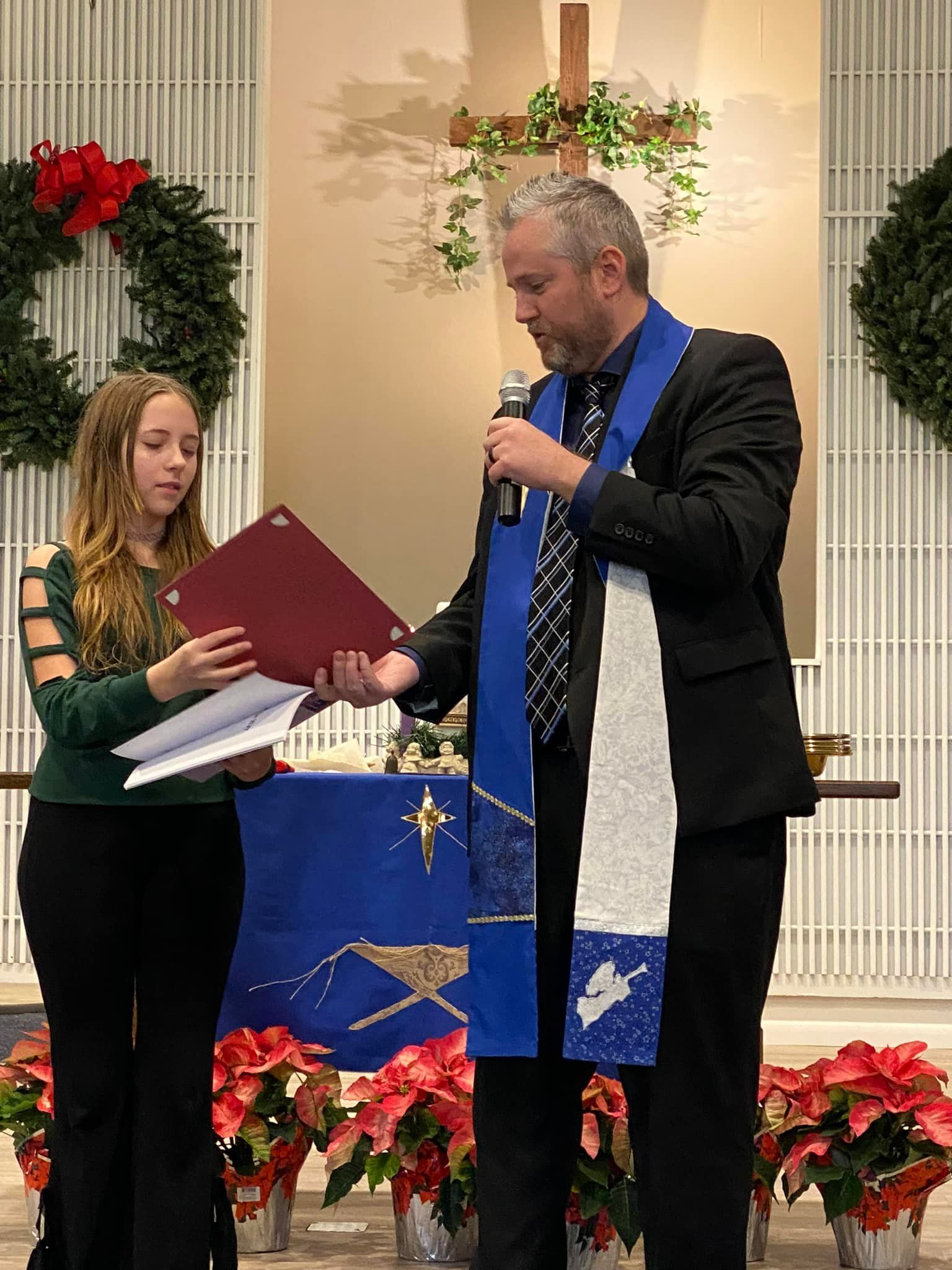 A man in clerical attire speaks into a microphone, while a young person holds a document at a church altar.