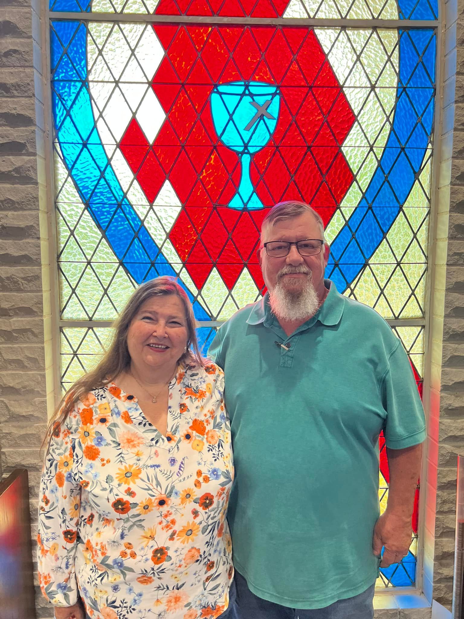 A couple smiles in front of a stained glass window depicting a chalice; red, blue, and yellow colors.