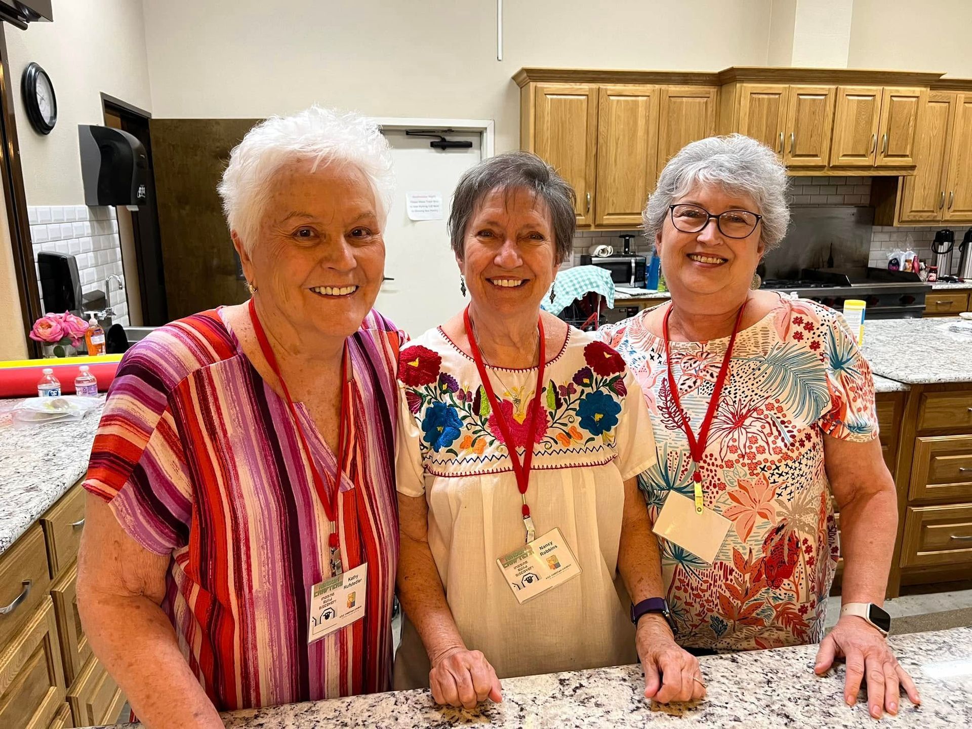 Three smiling women stand behind a counter in a kitchen. They wear name tags and colorful shirts.