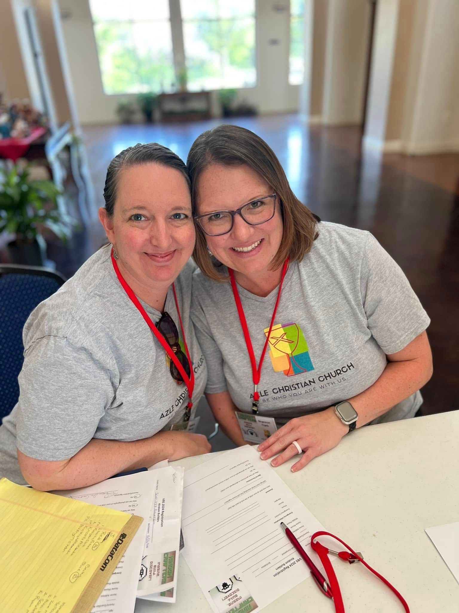 Two women smiling, sitting at a table with papers, wearing gray shirts and lanyards.