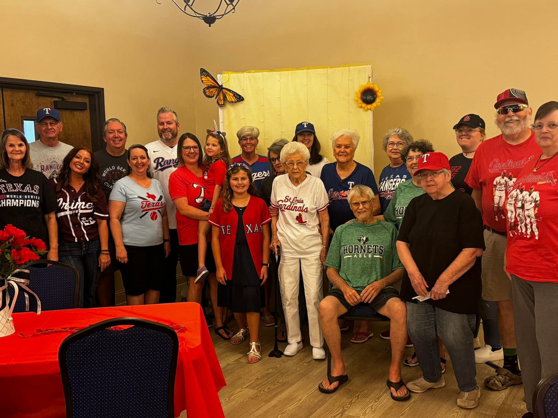 Group of people, mostly wearing red, posing for photo inside. Some are holding baseball caps.