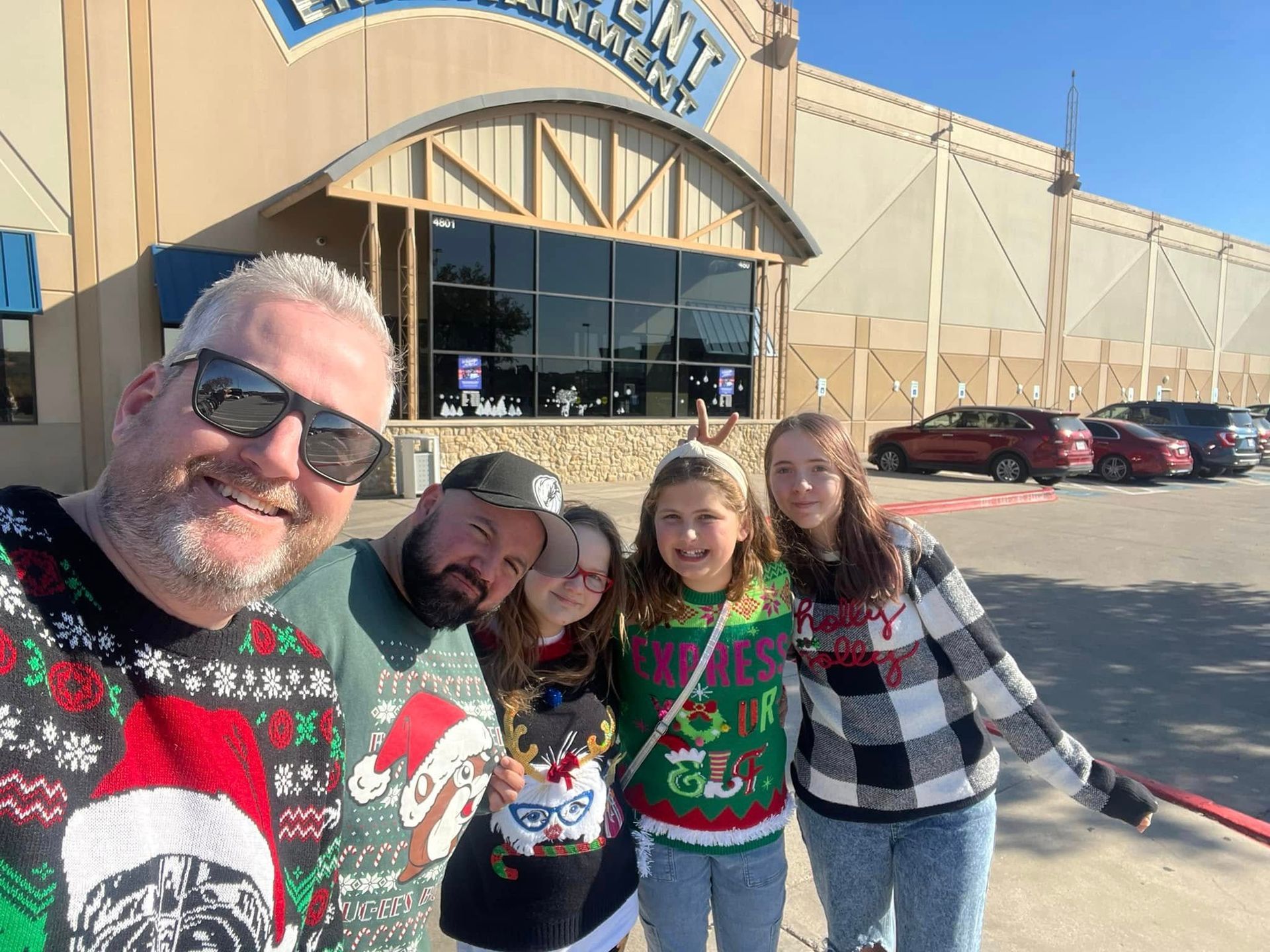 Family in Christmas sweaters in front of an entertainment center. Sun is shining.