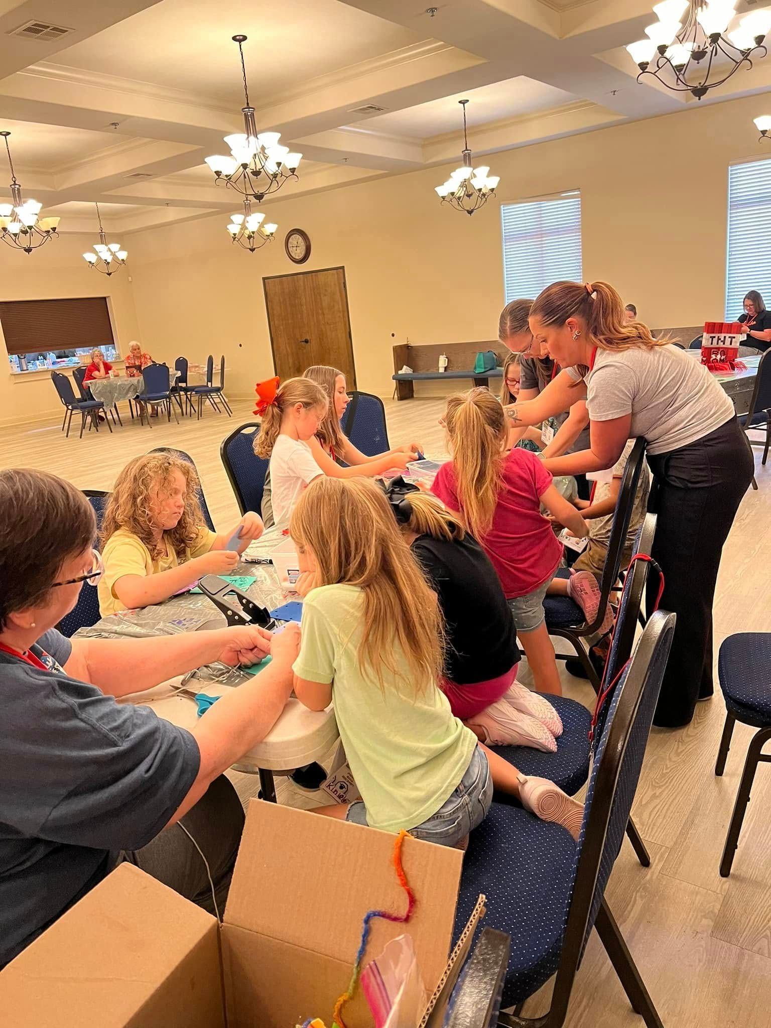 Children learning at a craft table; adults assisting. Bright room with chandeliers and round tables set.