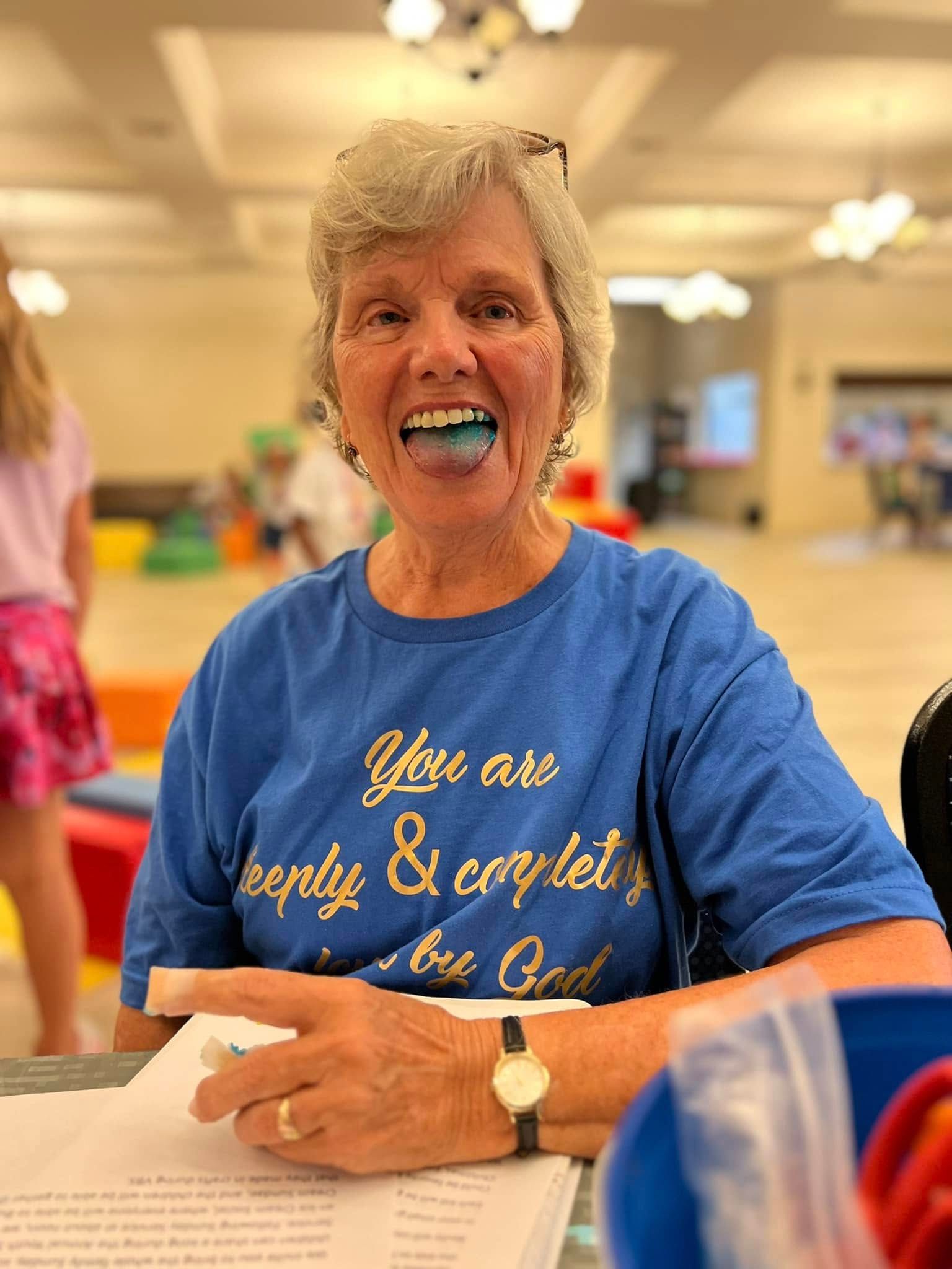 Woman smiling with blue candy in mouth, wearing a blue shirt with a gold inscription, indoors.