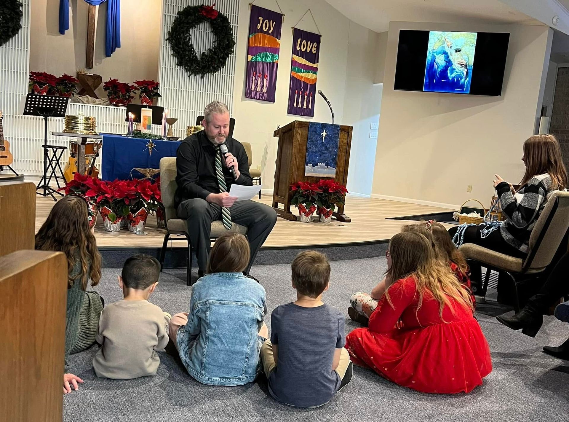 A man reads to a group of children in a church; stage decorated for Christmas.