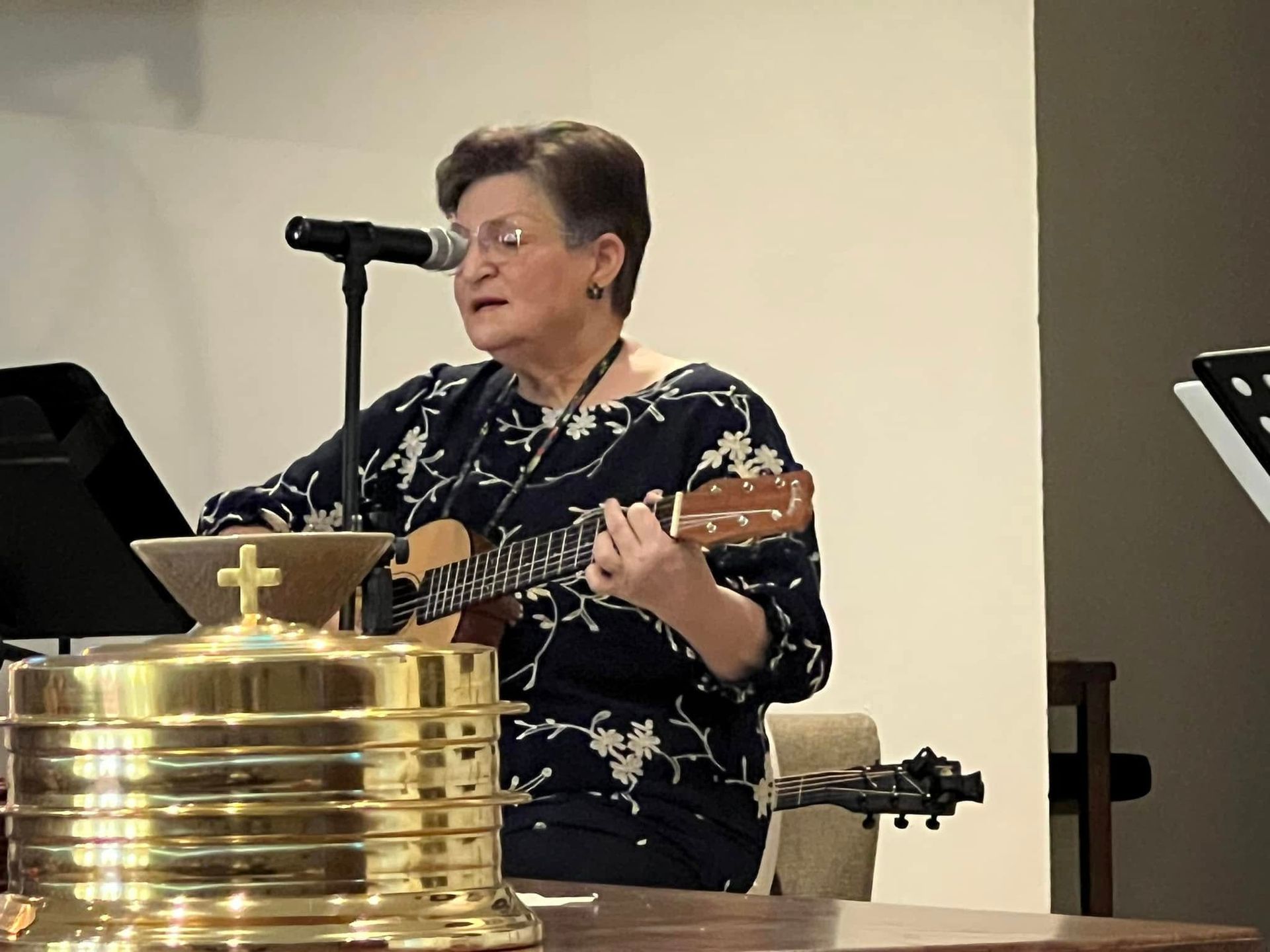 Woman playing ukulele and singing in front of microphone, standing near a communion set in a church.