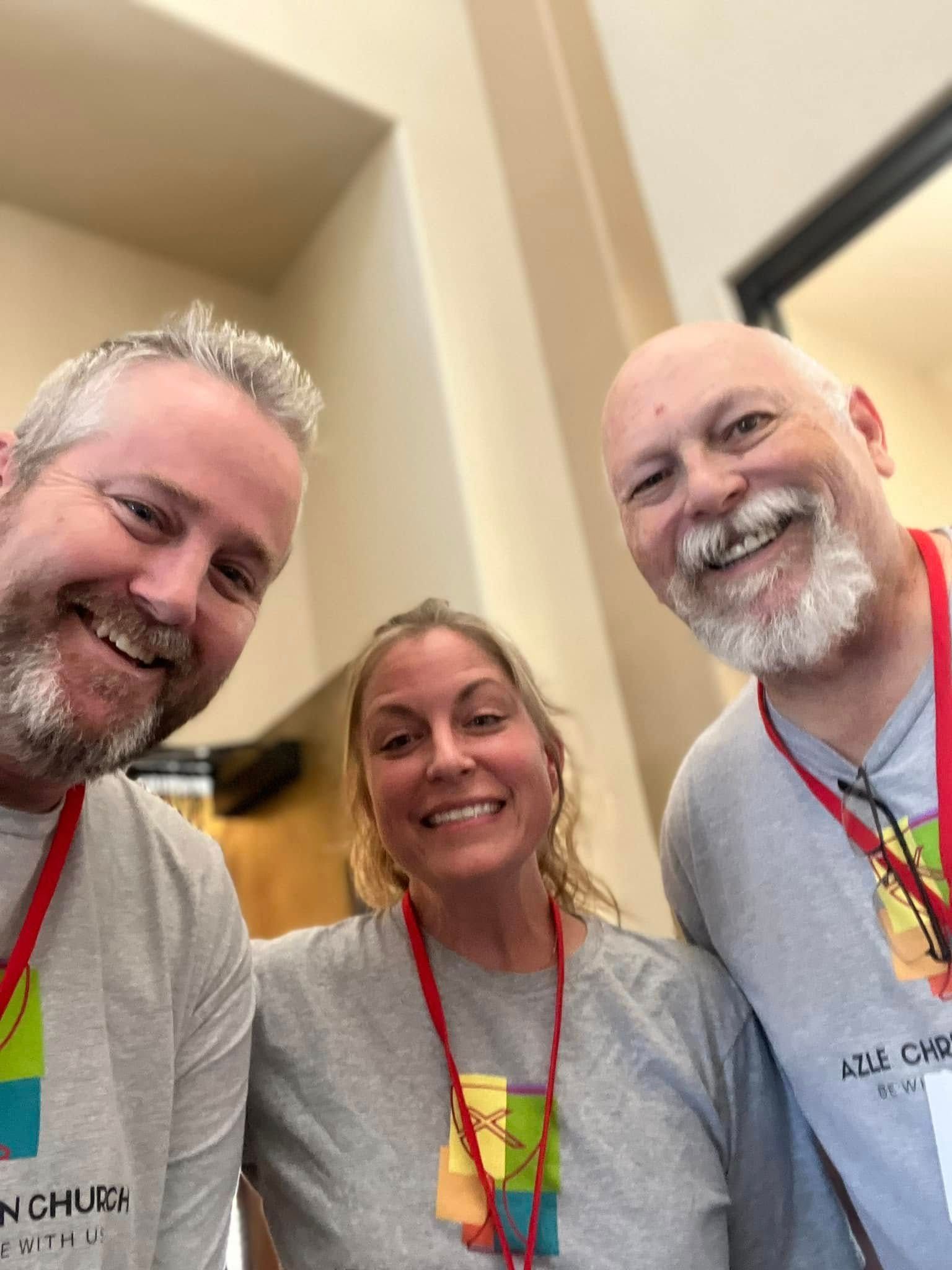 Three smiling people wearing gray shirts with red lanyards. Indoor setting, soft lighting.