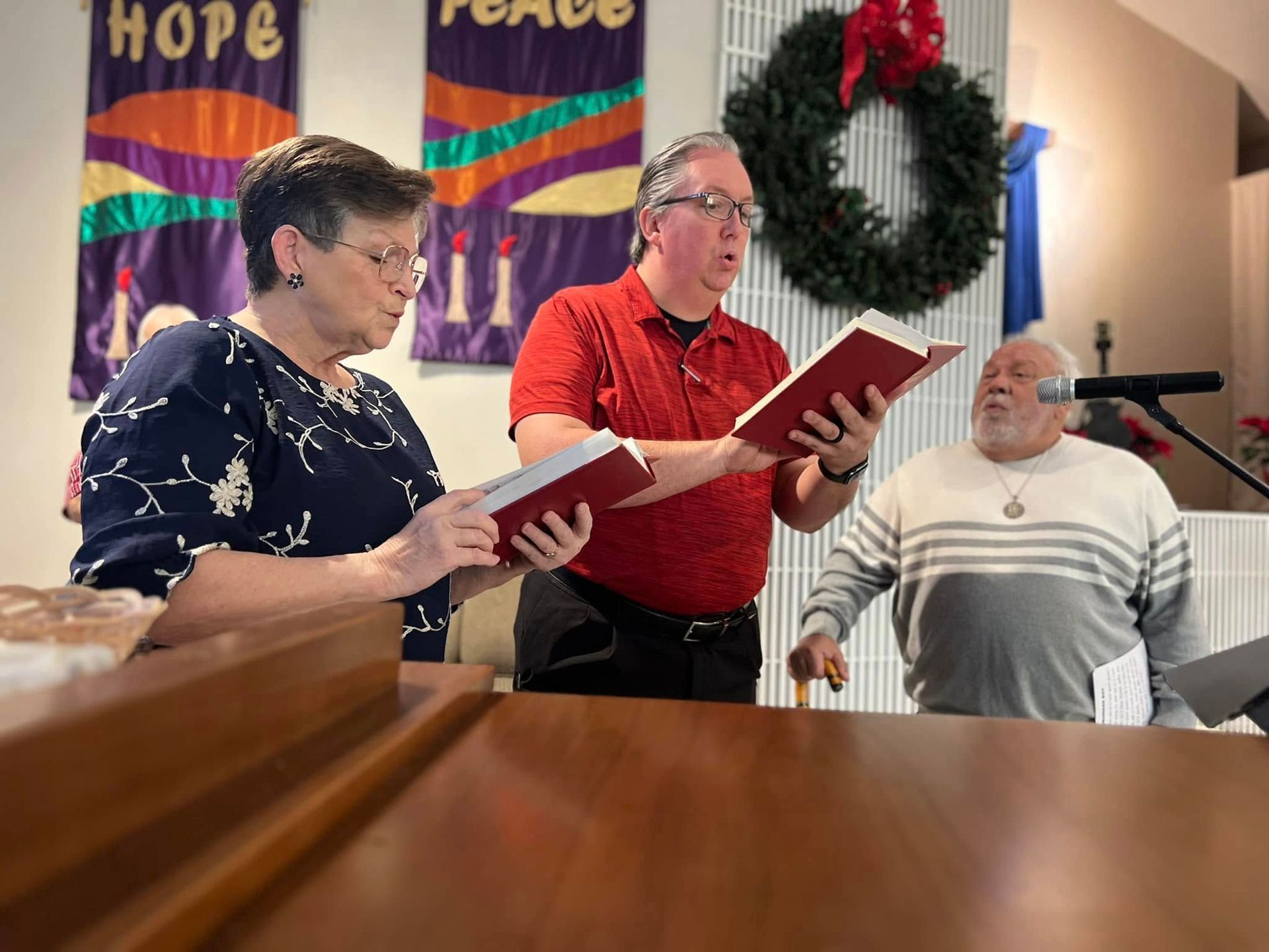 Three people singing in a church with open songbooks. A wreath and banners with the words 