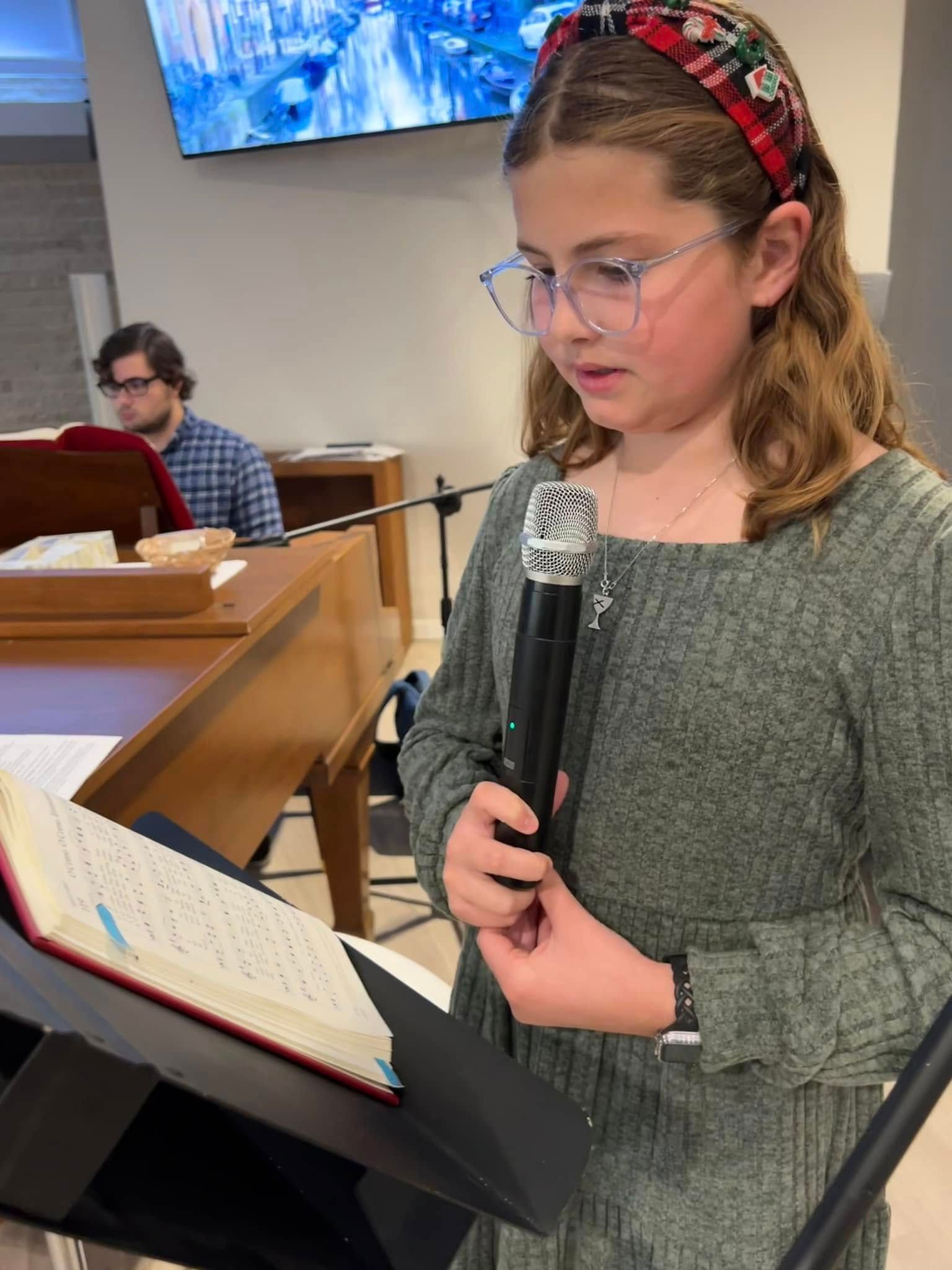 Girl holding microphone, reading from book at lectern. Man plays keyboard in background.