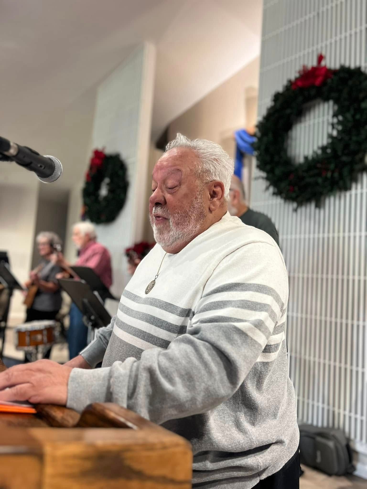 Man playing an instrument, singing, with microphone. Wreaths and musicians in the background.
