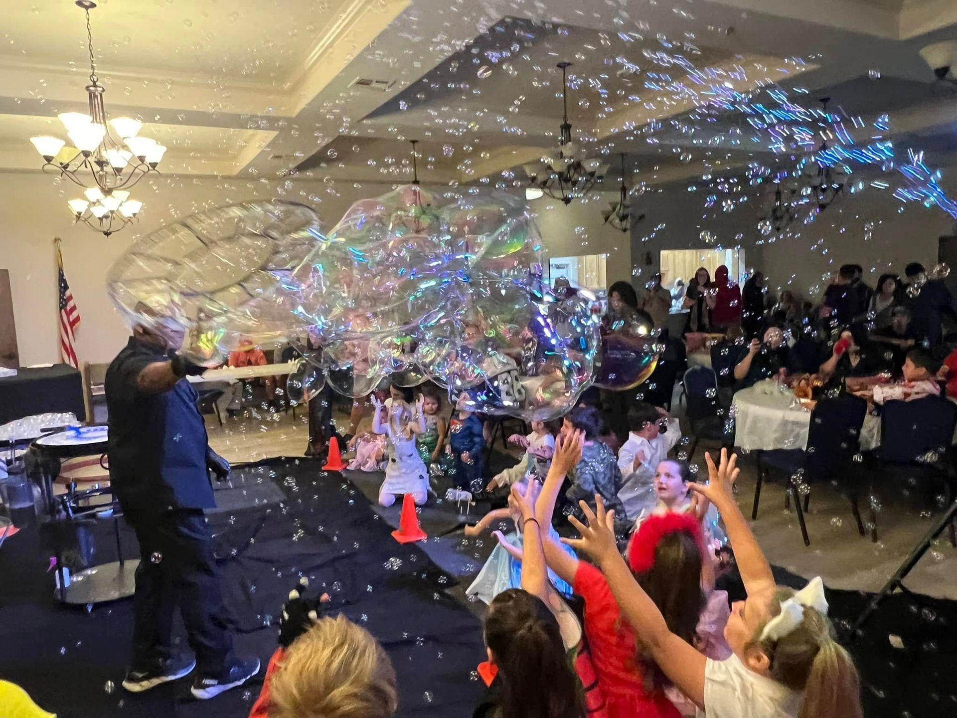 Magician makes giant bubbles as children reach up in an event hall.