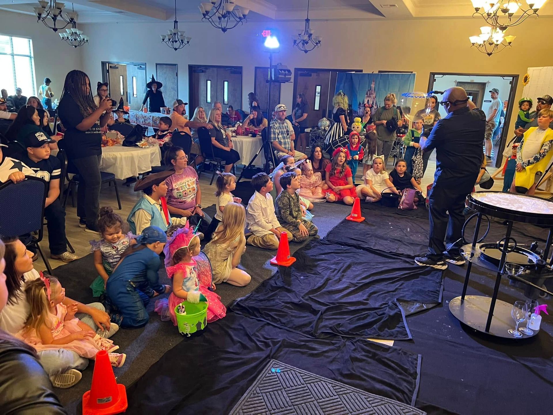Children in costumes watch a performer on a stage in a large room.