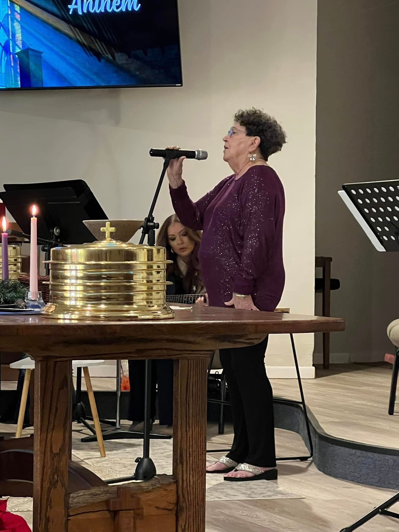 Woman singing into a microphone at a church service. There's a table with a chalice, and another woman playing the piano.
