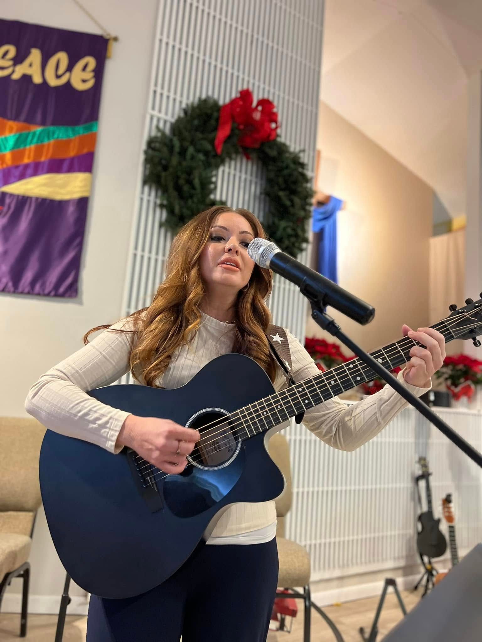 Woman playing a blue acoustic guitar and singing into a microphone. Wreath and banner in the background.