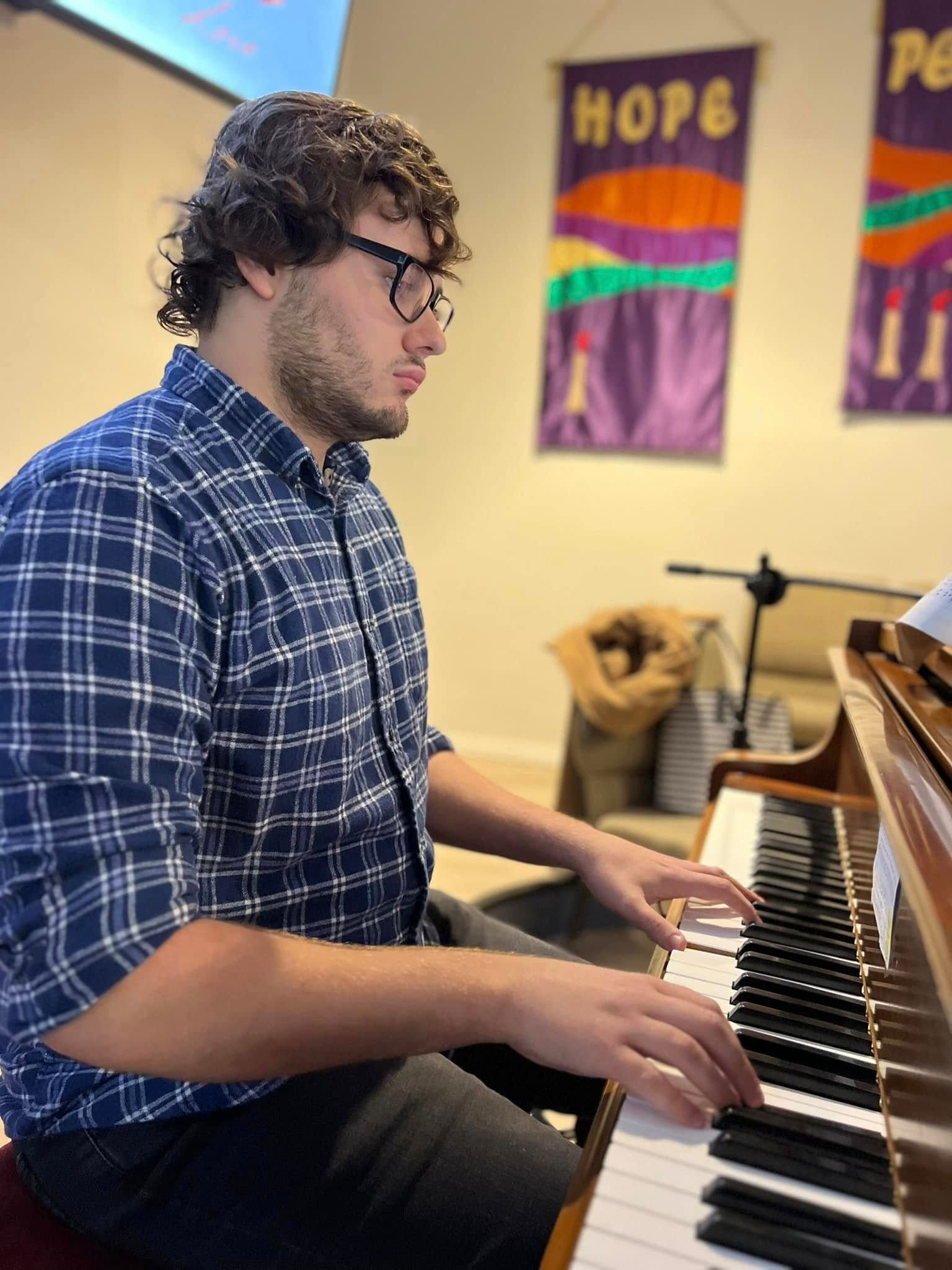 Man playing piano, wearing glasses and blue plaid shirt, in a room with banners and a microphone.
