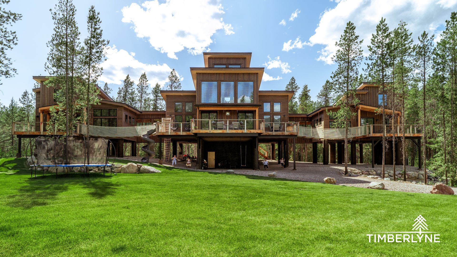 Modern multi-level timber home raised on stilts in a lush, wooded landscape under a sunny, blue sky.