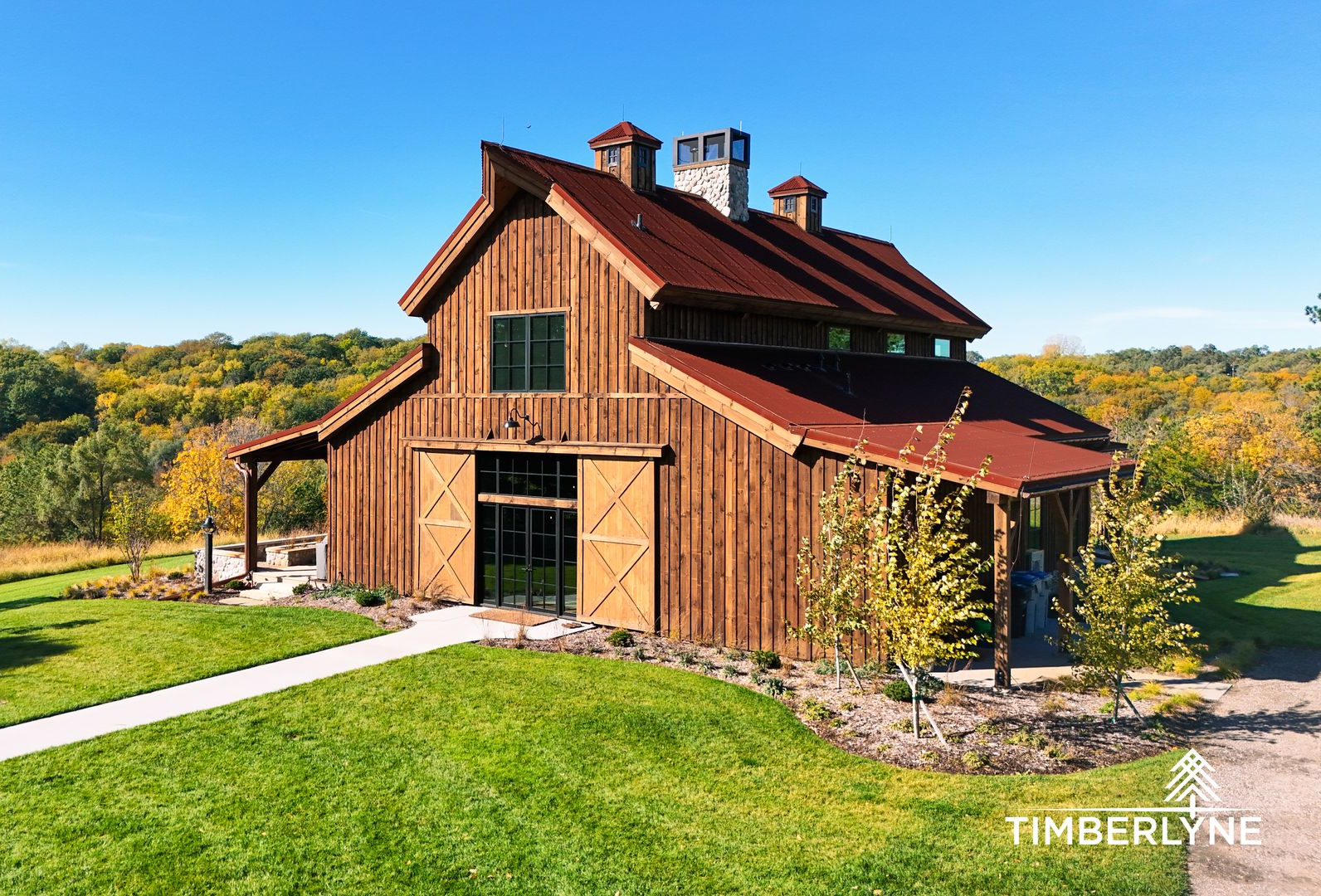 A rustic, wooden barn-style house with a dark metal roof sits on a grassy lawn under a clear blue sky.