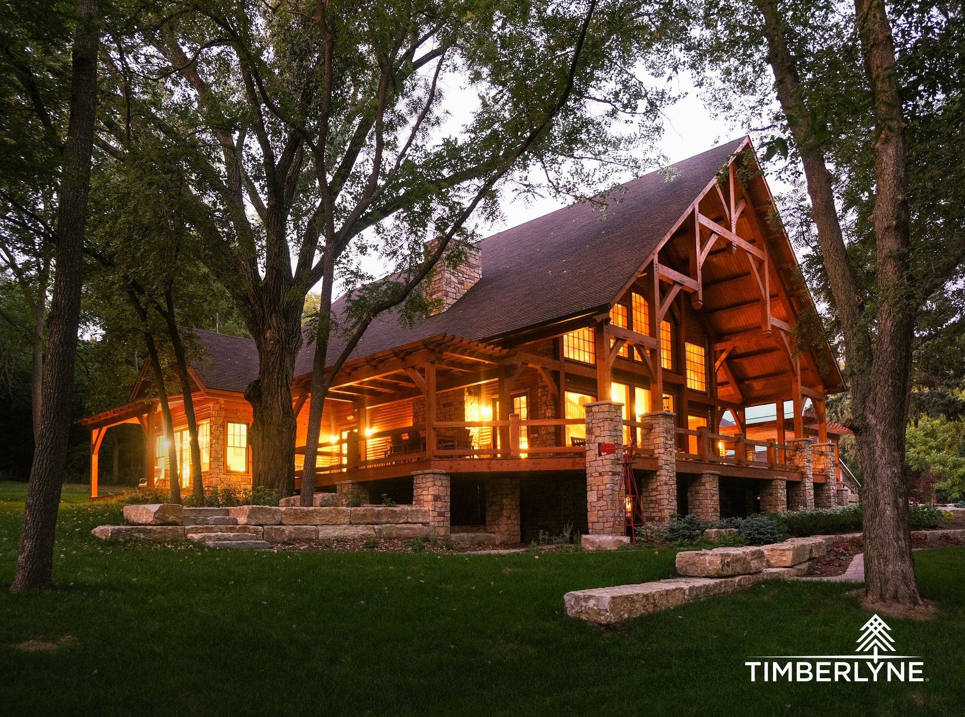 A timber-frame house with stone foundation and warm interior lighting glows at dusk amidst trees and landscaped lawn.