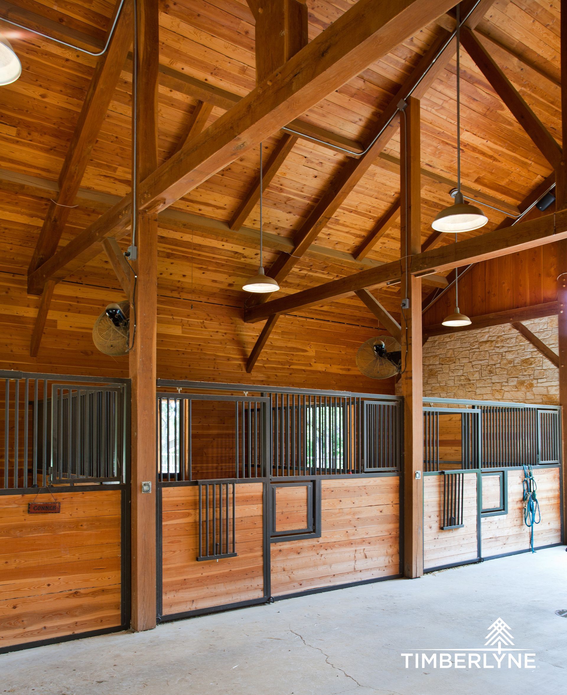 A rustic indoor horse stable with wood-paneled stalls, exposed timber beams, and pendant lighting.