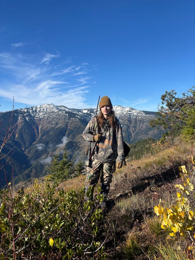 Hiker on a mountain trail with snowy peaks, blue sky, and yellow wildflowers.