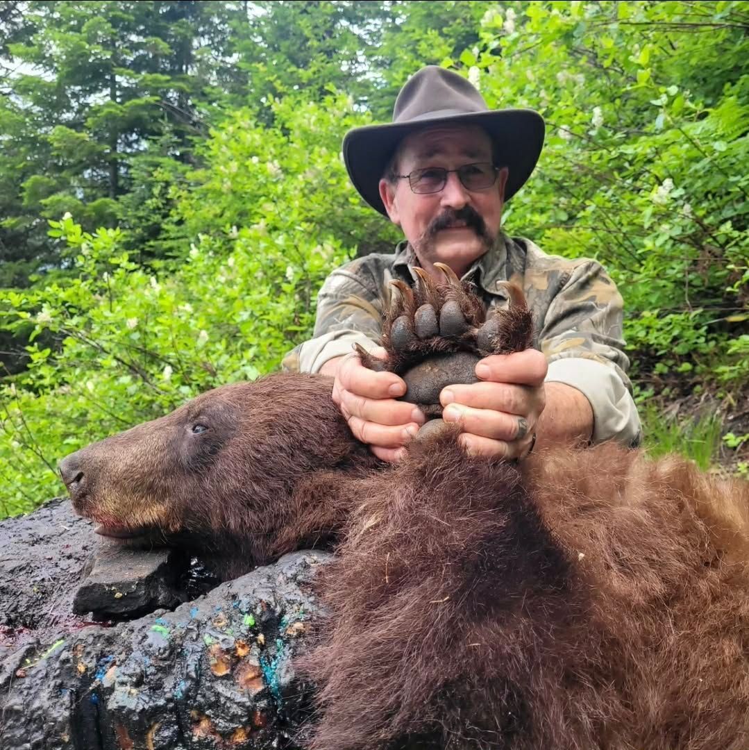 Hunter kneels beside a bear carcass, holding up a large paw in a green wooded area.