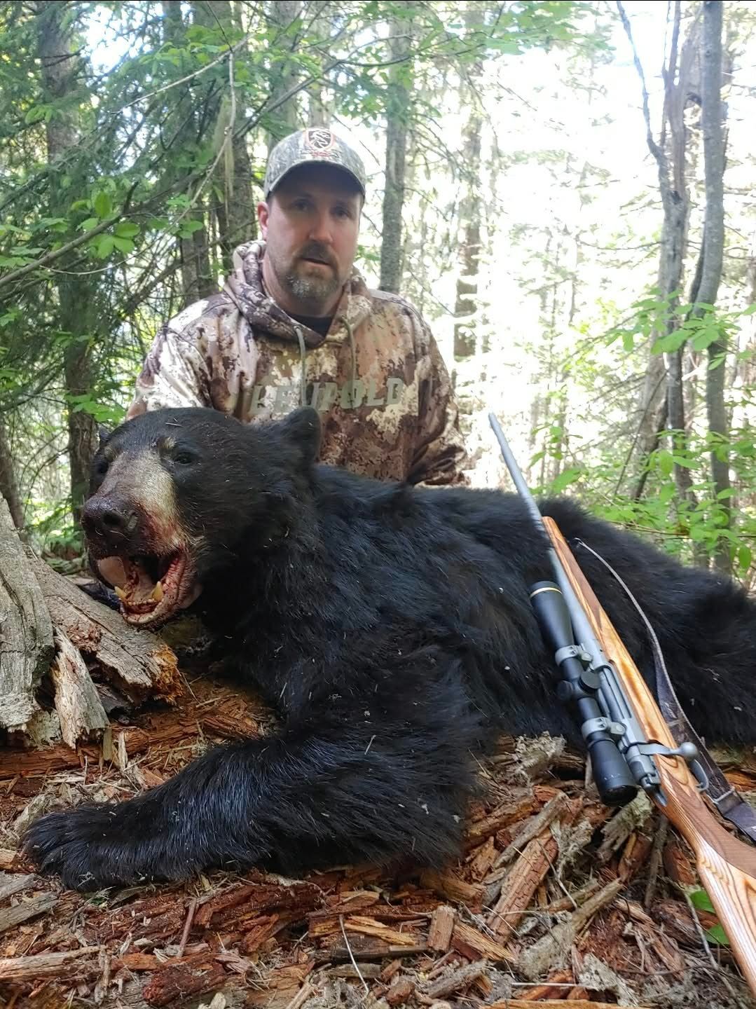 Hunter kneels beside a large black bear in a forest, with a rifle resting nearby.