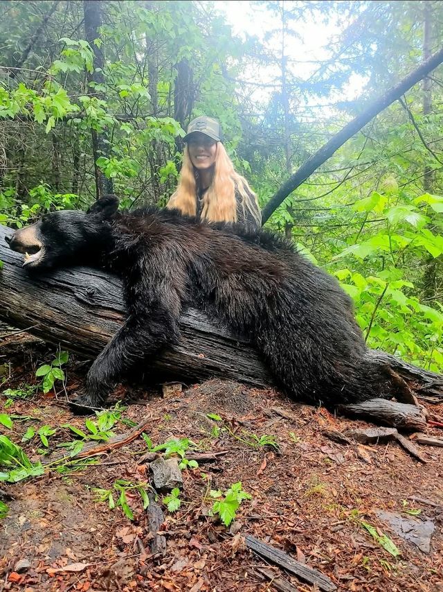 Hunter kneels behind a large dead bear in a muddy forest clearing.