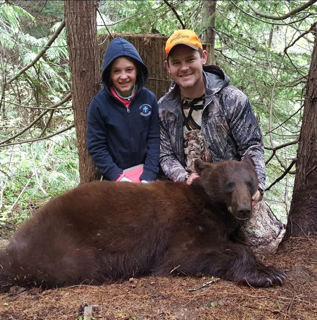 Two people in hoodies kneel beside a large brown bear in a wooded area.