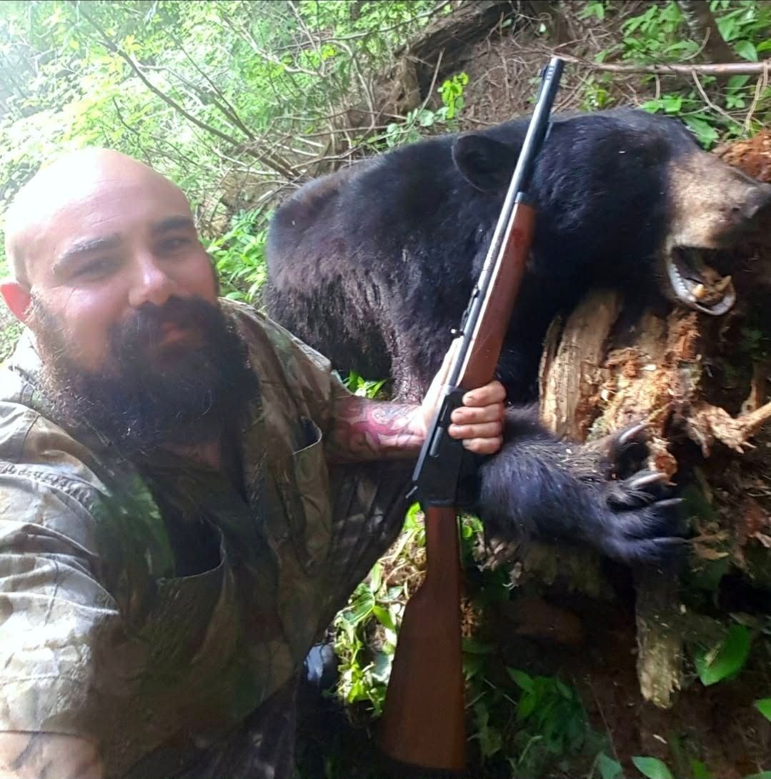 Man holding a rifle beside a dead bear in dense forest foliage